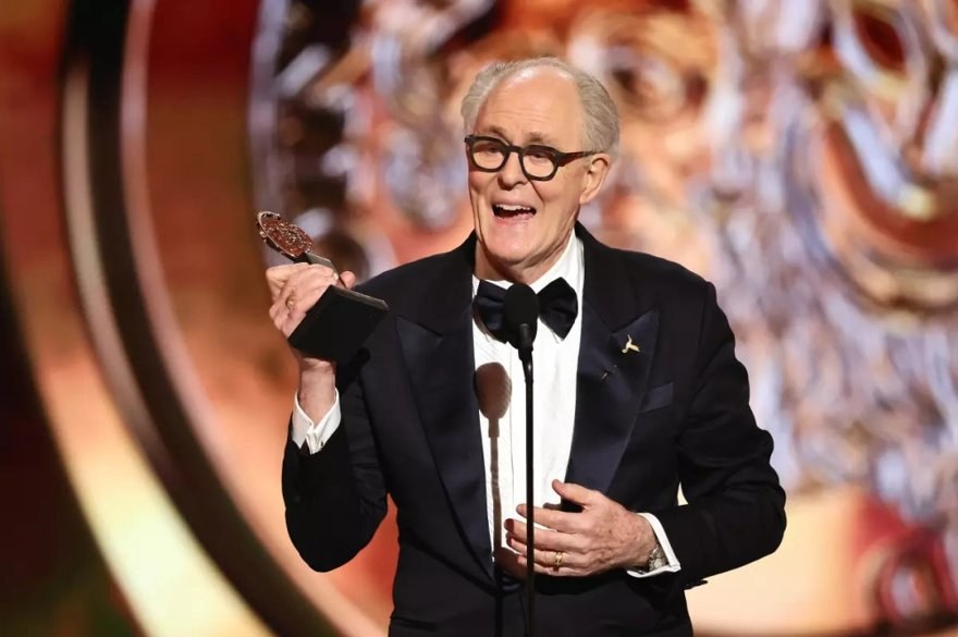 A single distinguished older man holding a silver award medallion under a spotlight.